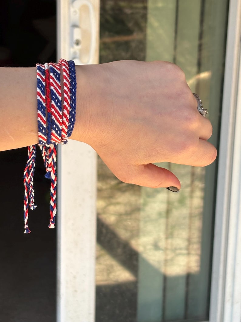 May include: A set of woven friendship bracelets in red, white, and blue. The bracelets are braided with a chevron pattern and tied with long, matching strands. The bracelets are worn on a wrist.