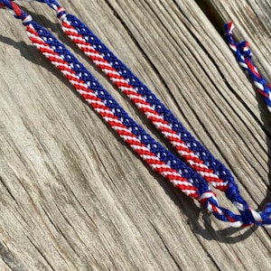 May include: Two friendship bracelets with a red, white, and blue pattern, resembling the American flag. The bracelets are made of woven thread and are displayed on a weathered wooden surface. The bracelets are a patriotic accessory.