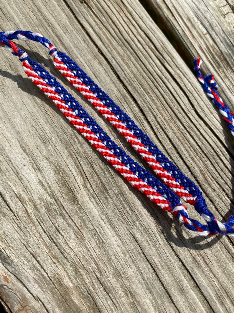 May include: Two friendship bracelets with a red, white, and blue pattern, resembling the American flag. The bracelets are made of woven thread and are displayed on a weathered wooden surface. The bracelets are a patriotic accessory.
