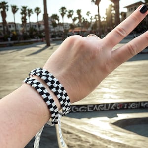 Könnte beinhalten: Zwei schwarz-weiß karierte Freundschaftsarmbänder werden am Handgelenk getragen. Die Armbänder bestehen aus gewebtem Faden. Der Hintergrund zeigt Palmen und einen sonnigen Himmel.