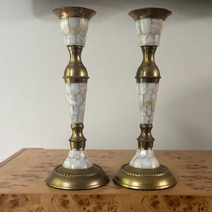 May include: A pair of matching candlestick holders featuring a combination of polished brass and iridescent white mother-of-pearl inlay. Each holder has a circular base, a slender stem, and a flared top, all accented with brass rings.