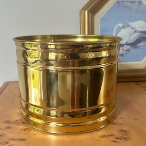 May include: A cylindrical brass planter with a polished, reflective gold-tone finish. The vessel features a smooth, unadorned surface with two subtle horizontal ridges encircling the body near the top and bottom edges, creating a simple, clean design.