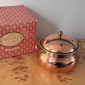 May include: A copper simmer pot with a matching lid featuring small ventilation holes and a brass knob. The pot has two brass side handles and is displayed next to its original red and white patterned box labeled Claire Burke Applejack & Peel.