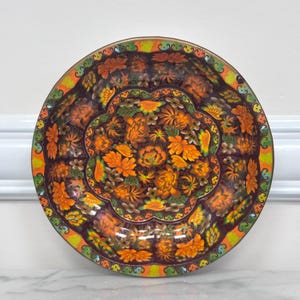 May include: A round decorative plate featuring a dark purple background with a vibrant floral pattern in shades of orange, yellow, and green. The rim is decorated with a repeating scalloped border in bright green, orange, and yellow tones.