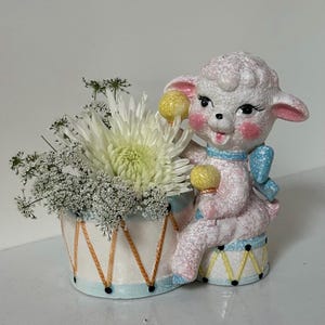 May include: A ceramic lamb figurine with a drum planter filled with white flowers. The lamb is pink with a blue bow and yellow drumsticks. The drum planter is white with a blue rim and gold accents.