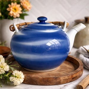 May include: A blue and white ceramic teapot with a bamboo handle and lid. The teapot is sitting on a round wooden tray. The teapot has a white spout and handle. Flowers are in the background.