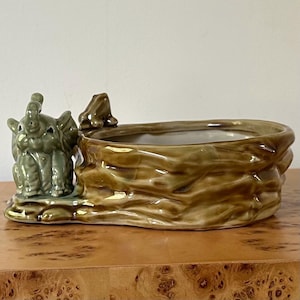 May include: A ceramic planter with a whimsical design. The planter features a light green elephant figurine on the left side and a textured, brown, tree-trunk-like container. The interior of the planter is white. The planter is sitting on a wooden surface.