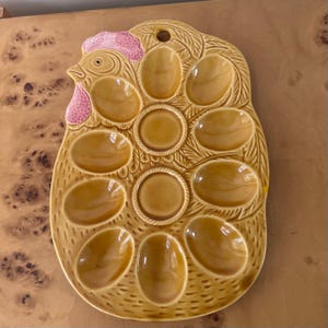May include: A glazed ceramic egg tray shaped like a chicken, featuring a mustard yellow finish with a pink comb and wattle. The tray includes ten oval indentations for eggs, a textured feather pattern, and a small circular hole at the top for hanging.