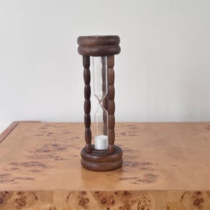 May include: A vertical hourglass featuring a dark brown wooden frame with a circular base and top connected by four turned spindles. A clear glass bulb is centered within the frame, containing white sand that is currently settled at the bottom.