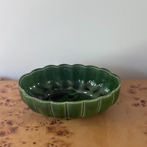 May include: An oval ceramic bowl with a deep forest green glaze and a scalloped, fluted edge. The interior features a smooth, reflective surface with subtle variations in color intensity, while the exterior has vertical ridges.