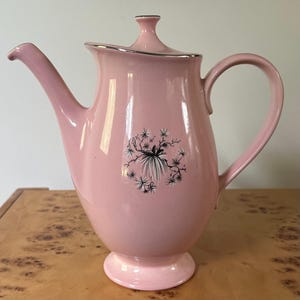 May include: A light pink ceramic teapot with a matching lid featuring a silver-toned rim. The body of the teapot has a curved handle, a long spout, and a pedestal base. A black and white botanical illustration of pine needles is centered on the front.