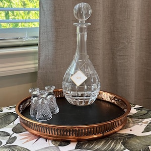 May include: A clear glass decanter with a faceted base and a matching stopper, featuring a diamond-shaped tag labeled GIN hanging from a gold chain, accompanied by three small matching crystal glasses on a round copper-toned metal serving tray.