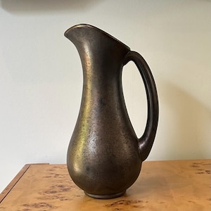 May include: A tall, slender pitcher with a rounded base and a curved handle. The surface features a mottled, metallic finish with blended tones of dark bronze, deep brown, and hints of muted gold, creating a textured appearance throughout the body.