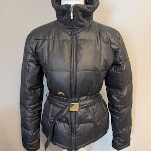 Puede incluir: Chaqueta acolchada negra con cuello alto y cinturón con hebilla dorada. La chaqueta es acolchada y tiene cierre de cremallera. La chaqueta se exhibe en un maniquí.