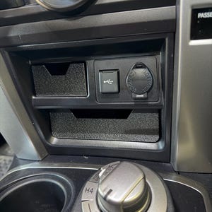 May include: A black plastic vehicle center console storage tray with a USB port and a 12V 120W power outlet. Below the tray is a silver rotary dial for four-wheel drive settings labeled H2, H4, and unlock, positioned near a cup holder.