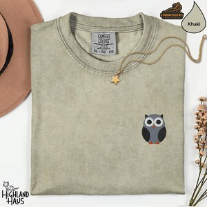 May include: Khaki-colored XL Comfort Colors t-shirt with an embroidered owl design. The owl is gray, black, white, and orange. A gold star pendant necklace is draped across the shirt. The Highland Haus logo is visible.