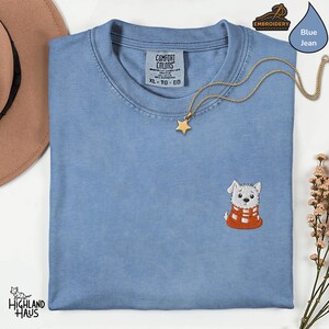 May include: A blue jean Comfort Colors t-shirt with an embroidered West Highland White Terrier wearing an orange scarf. A gold star pendant necklace rests on the shirt. The shirt is folded and ready to wear.