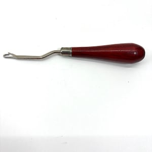 May include: A rug hooking tool featuring a smooth, dark reddish-brown wooden handle and a silver-colored metal shaft. The metal component includes a small, open hook at the tip, connected to the handle by a polished silver-toned metal ferrule.