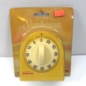 May include: A yellow Sunbeam kitchen timer in its original packaging. The circular timer features a cream-colored dial with black numbers from 0 to 55, a central rotating knob, and a red indicator arrow at the top. The packaging is yellow.