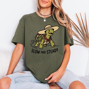 May include: Olive green t-shirt with a cartoon turtle wearing a hat and scarf. The turtle is walking on a path, with the words "SLOW and STEADY" printed below. The shirt is a comfortable, casual style.