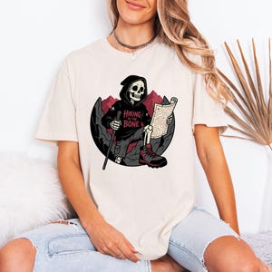 Pode incluir: Camiseta creme com um gráfico de um esqueleto em um moletom preto com capuz, lendo um mapa. O esqueleto está sentado com botas de caminhada e um bastão, com o texto "Hiking to the Bone". Montanhas vermelhas ao fundo.