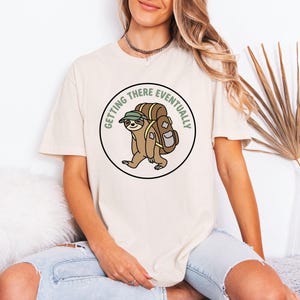 Puede incluir: Camiseta color crema con un gráfico circular de un perezoso con mochila y sombrero, con el texto "Getting There Eventually" en verde. El perezoso está representado en un estilo de dibujos animados, con colores marrón y beige. La camiseta tiene mangas cortas.