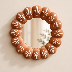 May include: A round mirror featuring a terracotta-colored frame with a scalloped, bubble-like edge. Each rounded section of the frame is decorated with a hand-painted white floral motif consisting of a central dot and radiating petal-like strokes.