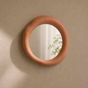 May include: A circular wall mirror featuring a thick, uneven, terracotta-colored frame with a matte, textured finish. The mirror glass is reflective and circular, set within the organic, hand-molded appearance of the clay-like border.