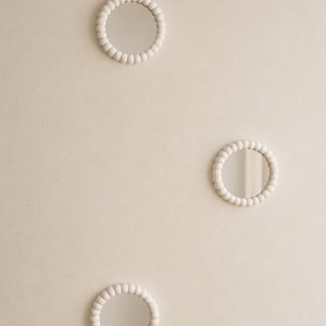 May include: Three circular wall mirrors, each featuring a frame composed of a continuous row of small, matte off-white spheres. The mirrors are arranged in a vertical, staggered formation, reflecting light on their smooth, clear glass surfaces.