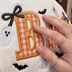 May include: A cream-coloured fabric featuring an embroidered orange and white gingham letter B with a black bow accent. The design includes small white ghost figures and black bat silhouettes embroidered around the letter on the textile surface.