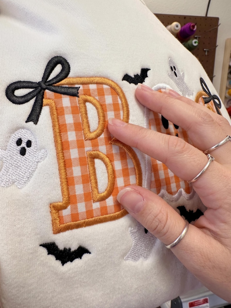 May include: A cream-coloured fabric featuring an embroidered orange and white gingham letter B with a black bow accent. The design includes small white ghost figures and black bat silhouettes embroidered around the letter on the textile surface.