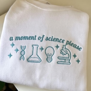 May include: A white crewneck sweatshirt featuring teal embroidery that reads "a moment of science please" above a row of icons including a DNA strand, a laboratory flask, a lightbulb, and a microscope, all accented with small four-pointed stars.