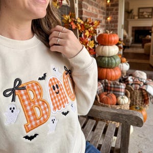 May include: A cream-coloured crewneck sweatshirt featuring the word BOO embroidered in an orange and white gingham pattern. The design includes small white ghost figures and black bats, with a black bow accent on the letter B.