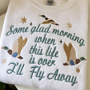 May include: A white crewneck sweatshirt featuring embroidered mallard ducks in flight and gold star accents. The text reads, "Some glad morning when this life is over I'll Fly Away" in dark green cursive and block lettering, centred on the chest.