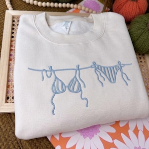 Può includere: Una felpa girocollo color crema con un ricamo azzurro che raffigura un top bikini e uno slip coordinato appesi a un filo per il bucato. Il costume da bagno ricamato presenta un sottile motivo a righe e lacci visibili sui lati.