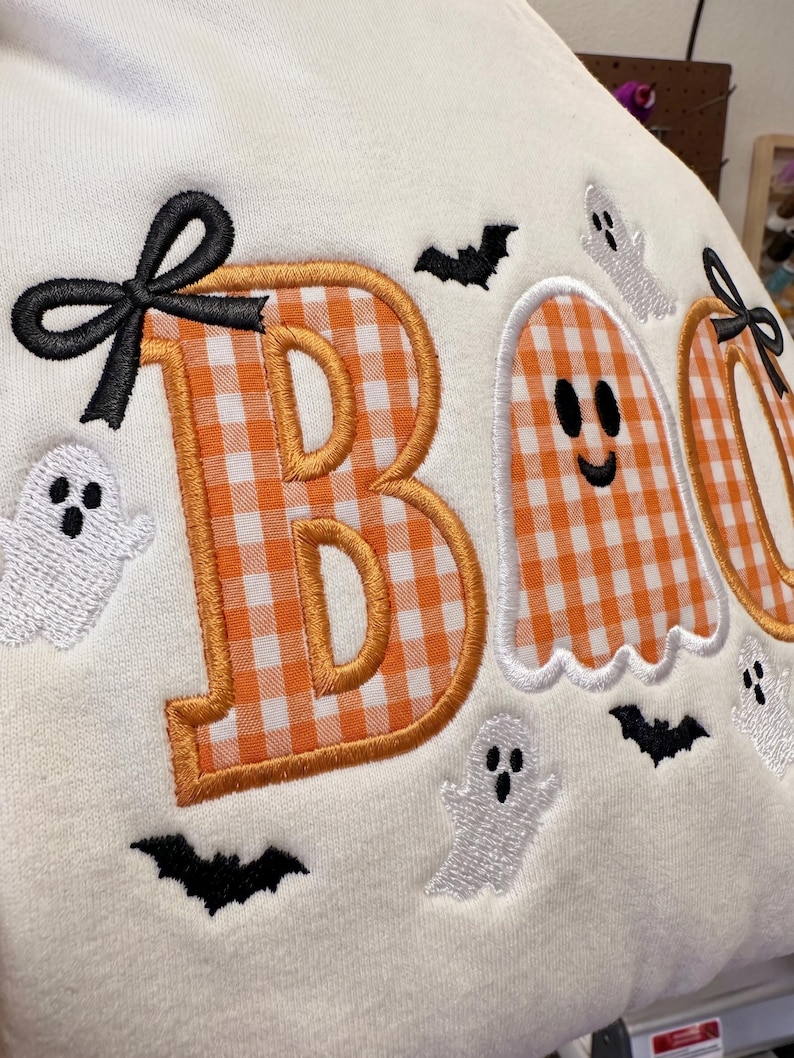 May include: A cream-coloured fabric featuring an embroidered design with the word BOO. The letter B is orange and white gingham with a black bow. The letter O is a white ghost with a smiling face. Small white ghosts and black bats are scattered.