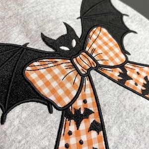 May include: A light gray sweatshirt featuring an embroidered design of a black bat with white eyes centered behind a large orange and white gingham bow. The bow has long tails decorated with small black bat silhouettes and black dots.