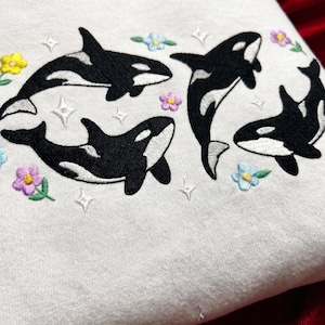Peut inclure: Un sweat-shirt blanc orné d'une broderie représentant trois orques noires et blanches en train de nager. La broderie comprend de petites fleurs colorées jaunes, bleues et violettes, ainsi que des accents en forme d'étoiles blanches dispersés.
