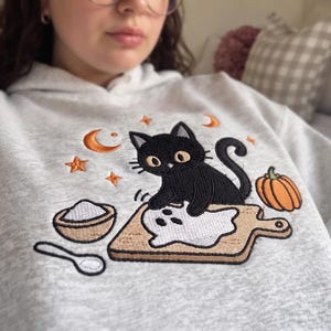 May include: A light gray hooded sweatshirt featuring an embroidered design of a black cat on a wooden cutting board with a white ghost shape, a small bowl of white powder, a spoon, an orange pumpkin, and orange stars and crescent moons.
