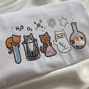 Puede incluir: Una sudadera blanca con un diseño bordado de cinco gatos interactuando con material de laboratorio y símbolos científicos. El bordado incluye las fórmulas químicas H2O y O2, una molécula, un átomo y una estructura de anillo de benceno.