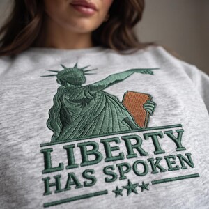 May include: A light grey heather sweatshirt featuring an embroidered design of the Statue of Liberty in dark green with a copper-coloured tablet. Below the figure, the text reads LIBERTY HAS SPOKEN in dark green letters with three stars.