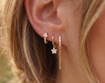 May include: Three gold hoop earrings worn in a row. The first is a small hoop with a three-stone cluster. The second is a plain hoop with a dangling flower charm featuring clear stones. The third is a hoop with a vertical row of stones and a chain.