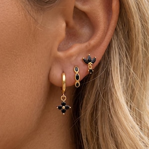 May include: A set of three gold-toned earrings featuring black stones. The lowest earring is a small hoop with a four-petal flower charm, the middle is a vertical marquise-shaped stud, and the top is a three-stone fan-shaped stud earring.