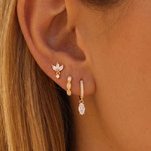 Pode incluir: Três brincos dourados usados numa única orelha. O brinco superior é um pino em forma de flor com pedras de corte marquise. O brinco do meio é uma pequena argola com três pedras marquise. O inferior é uma argola com uma pedra pendente.
