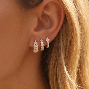 Peut inclure: Trois boucles d'oreilles créoles en or portées en rangée sur le lobe de l'oreille. La première présente un motif de feuilles verticales avec de petites pierres claires. La deuxième a un motif texturé de pierres empilées. La troisième est une créole simple avec une breloque en forme de trèfle.