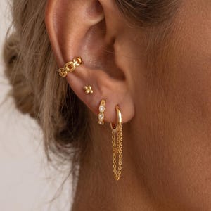 Può includere: Un ear cuff dorato con design a maglie testurizzate, un piccolo orecchino a lobo con quattro punti, un orecchino a cerchio dorato con tre pietre chiare incastonate e un orecchino a cerchio dorato con due delicate catenelle pendenti alla base.