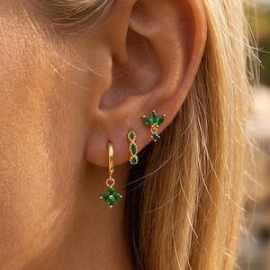 May include: Three gold-tone earrings featuring green marquise-cut stones. One earring is a small hoop with a dangling four-petal flower charm, one is a vertical bar with two stones, and one is a stud with a three-petal leaf design.