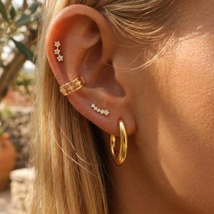 Puede incluir: Una oreja está adornada con una colección de pendientes dorados. El conjunto incluye un pendiente de aro, un ear cuff en forma de estrella y múltiples pendientes de botón con diseños de estrella y lineales. Los pendientes están engastados con pequeñas piedras preciosas transparentes.