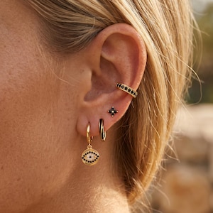 May include: Close-up of a person's ear adorned with gold earrings and ear cuffs. The ear features a gold ear cuff with black stones, a small black stud earring, a black and gold hoop earring, and a gold hoop earring with an eye pendant.