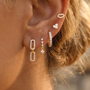 May include: A collection of gold earrings, including a pair of linked oval studs, a dangling earring with a square gem, and a small hoop earring. Additional earrings include a small lip-shaped stud and a heart-shaped stud, all encrusted with small, sparkling stones.